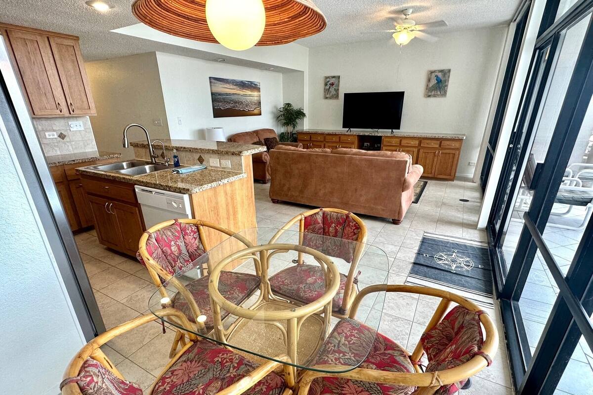 A modern kitchen with wooden cabinets, a dining area with wicker chairs and a glass table, a living room with a sofa, and a large window offering a view.