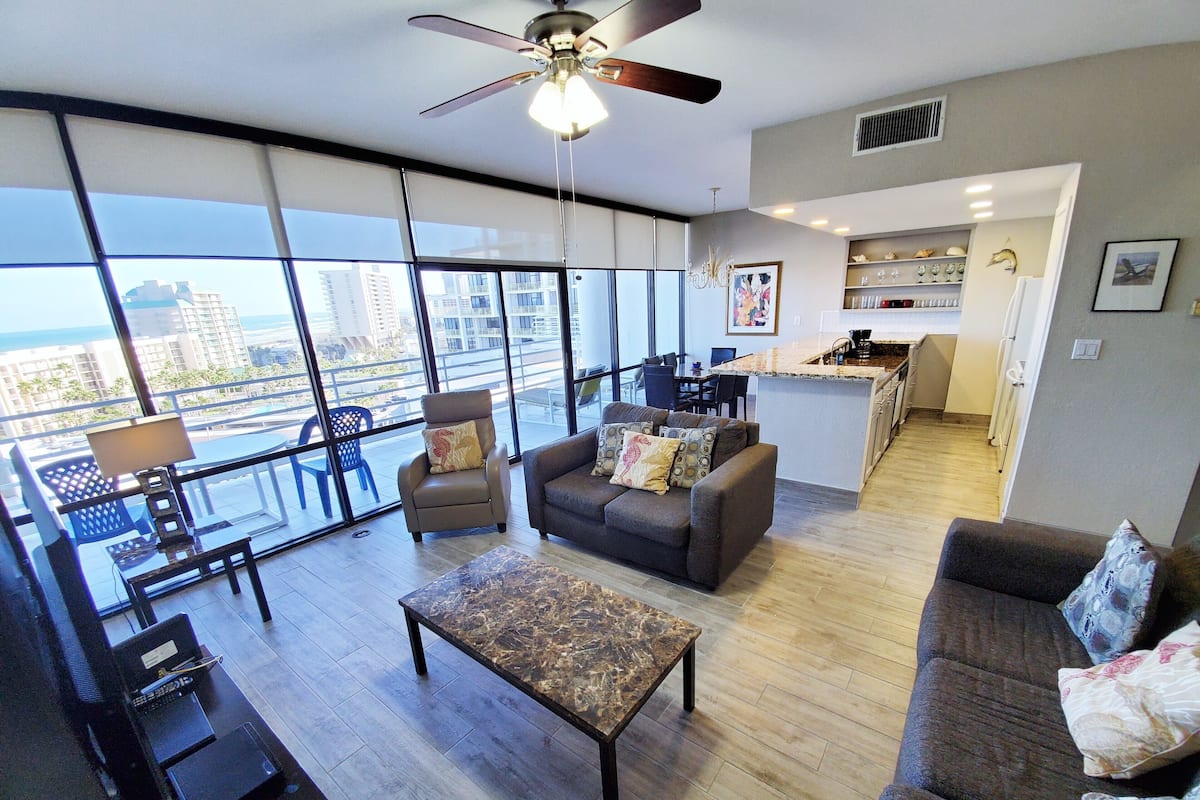 A spacious living room with a large window, a ceiling fan, a sofa, a coffee table, and a small kitchen area.