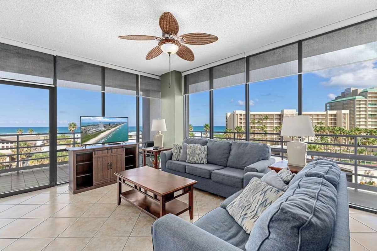 A spacious living area with a large gray sofa, a wooden coffee table, and a television on a stand. There is a ceiling fan, large windows offering a view of the city, and a balcony with a railing.