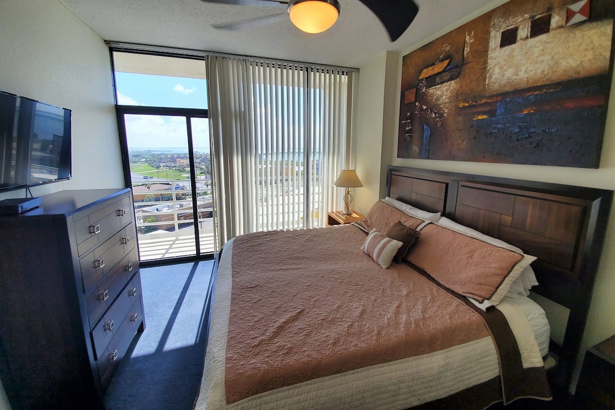 A hotel room with a bed, a dresser, a TV, a ceiling fan, a window with curtains, and a painting on the wall.