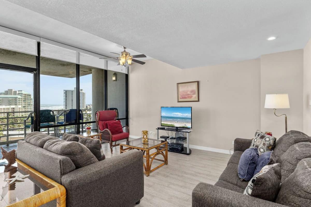 A modern living room with a large window, a ceiling fan, a flat-screen TV, and a balcony with a view of the city.