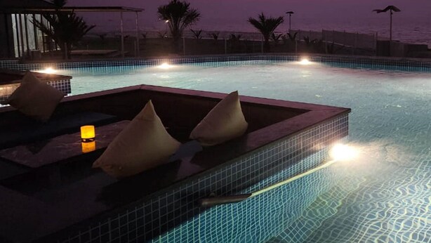 An infinity pool with a view of the ocean at dusk, equipped with lounge chairs and a small table with a candle. - Casa De Turtle  (Shrivardhan)