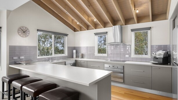A modern kitchen with a central island, grey cabinetry, and a stainless steel range hood. There are two windows providing natural light and a clock on the wall.
