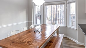 Comedor con mesa de madera, sillas blancas, lámpara colgante y ventanas amplias con vista a árboles.