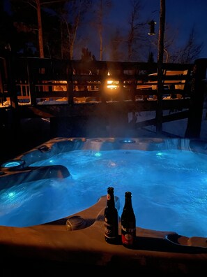 Jacuzzi con dos botellas de cerveza, rodeado por una terraza de madera y árboles.