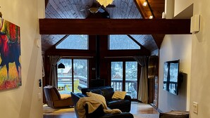 Amplia sala de estar con techo alto, vigas de madera, un gran candelabro, un sofá de cuero, un televisor y vistas a un paisaje nevado a través de amplios ventanales.