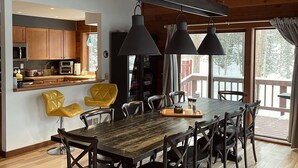 Una cocina moderna con armarios de madera, un comedor con una mesa grande y sillas, y vistas al exterior a través de amplias ventanas.