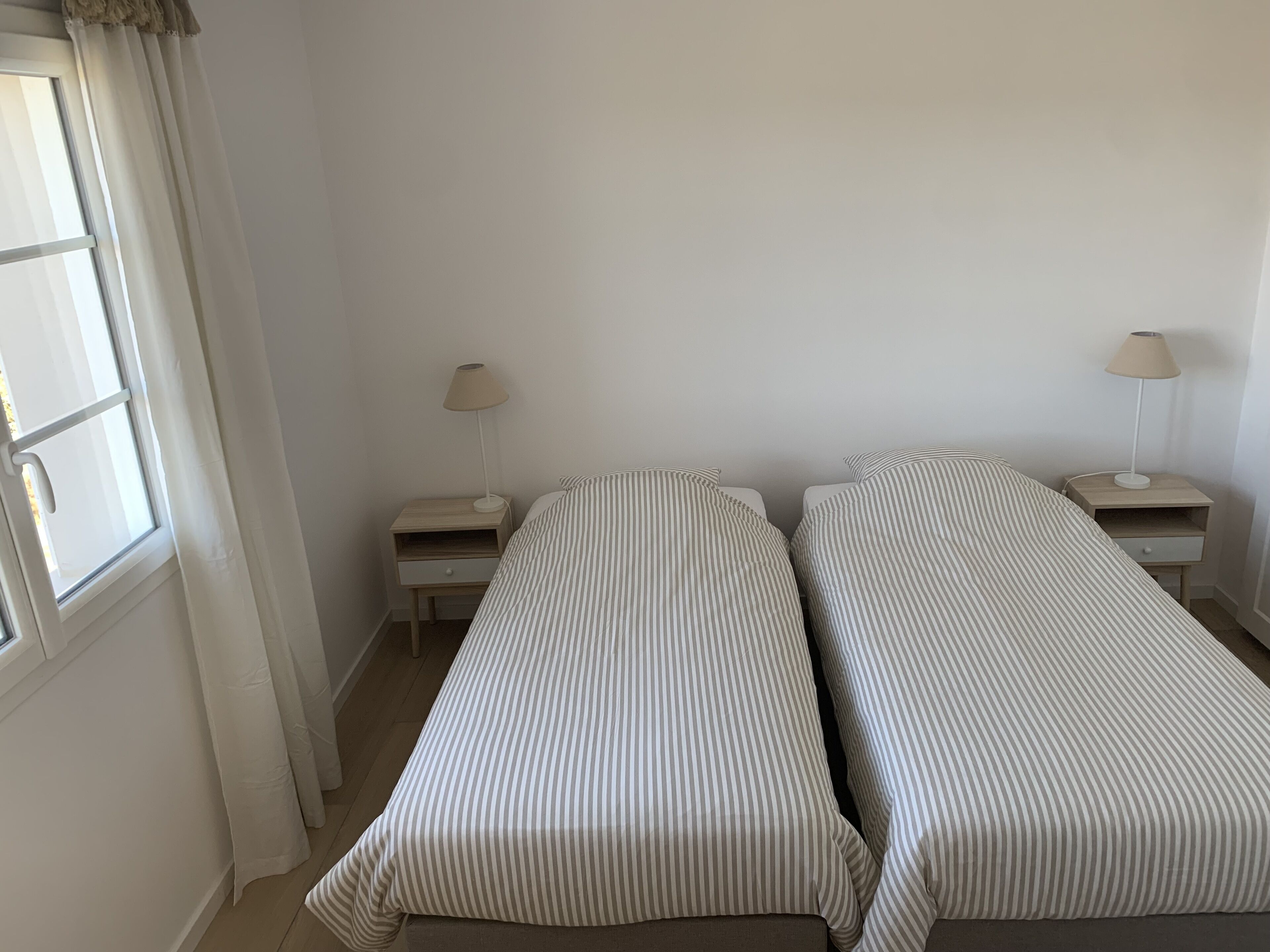 Habitación sencilla y limpia con dos camas individuales, una ventana con cortinas y dos mesitas de noche con lámparas.