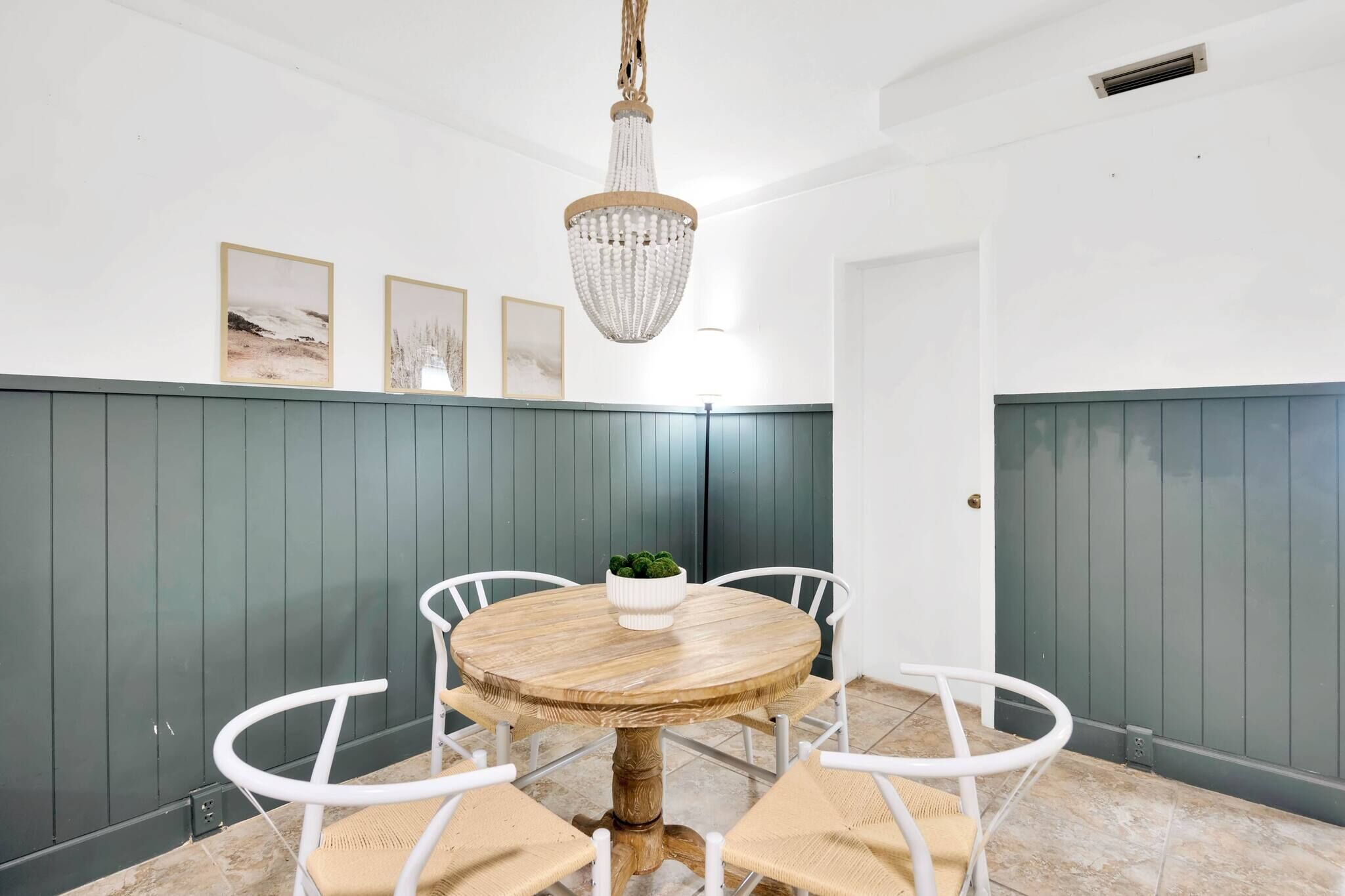 A dining area with a round wooden table, white chairs, a chandelier, and a green wall with vertical paneling.