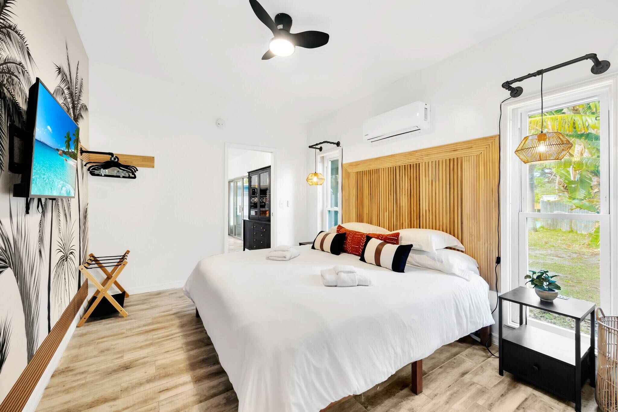 A hotel room with a large bed, a TV mounted on the wall, a ceiling fan, and a window with a view of greenery.