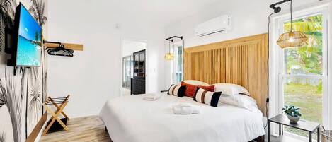 A hotel room with a large bed, a TV mounted on the wall, a ceiling fan, and a window with a view of greenery.