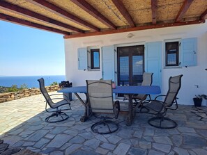 A covered outdoor dining area with a table and chairs, a stone floor, and a view of the sea.