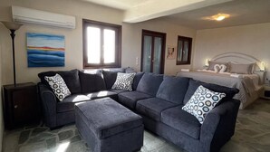 A living room with a sectional sofa, ottoman, stone flooring, air conditioning unit, windows, and artwork. - Dorothea Panoramic Villa.One of a kind, a hidden gem on the isle of Paros.Helios (Paros)