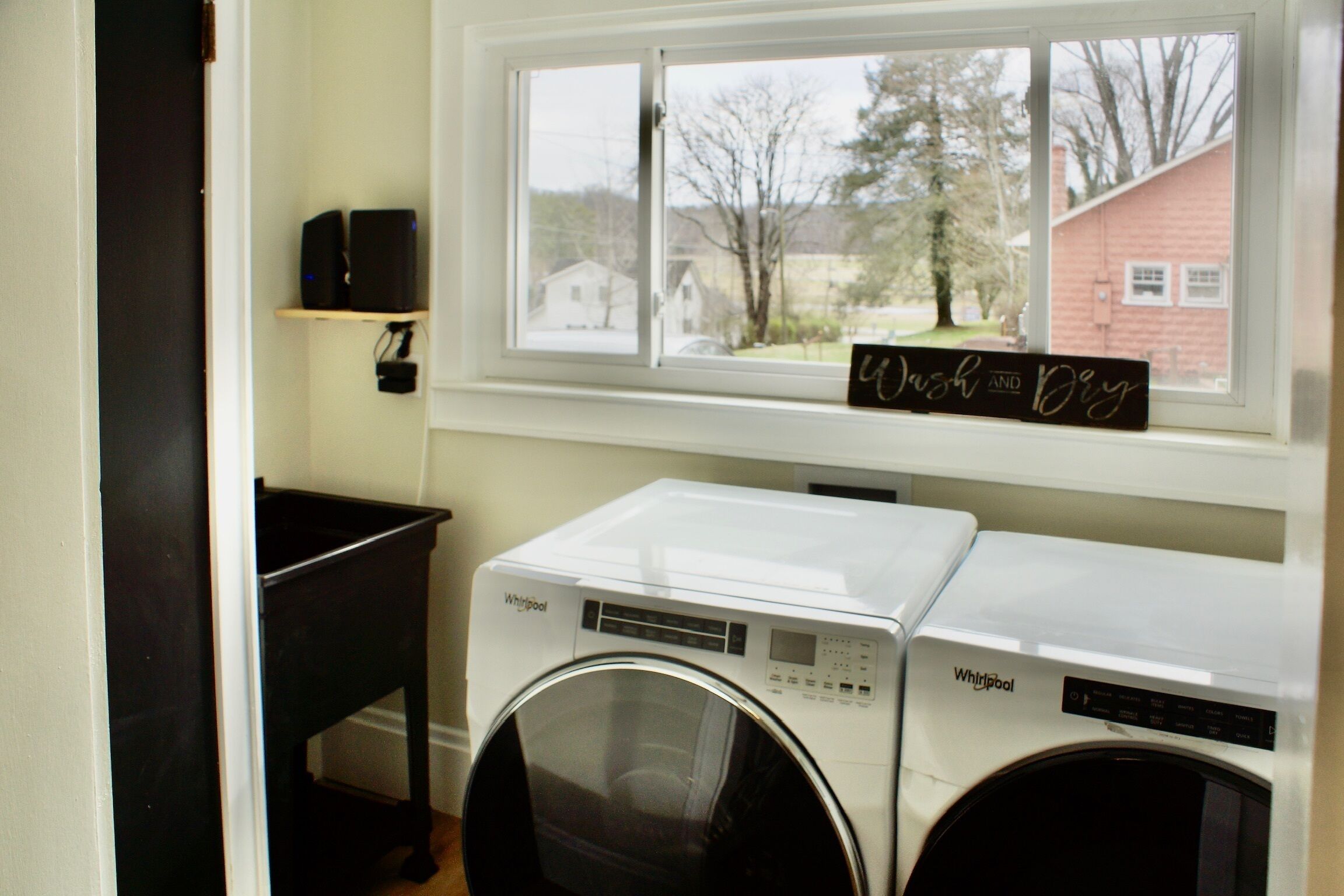 Una sala de lavandería con dos lavadoras y secadoras Whirlpool, un estante negro con altavoces y un ventanal con vistas a casas.