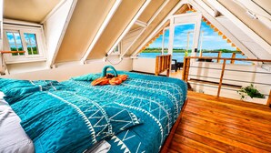 A bedroom with a large bed, wooden flooring, and a view of a body of water through a window. - The Sea Spot! Hip-Nautic Floating Lodge (Bocas del Toro)