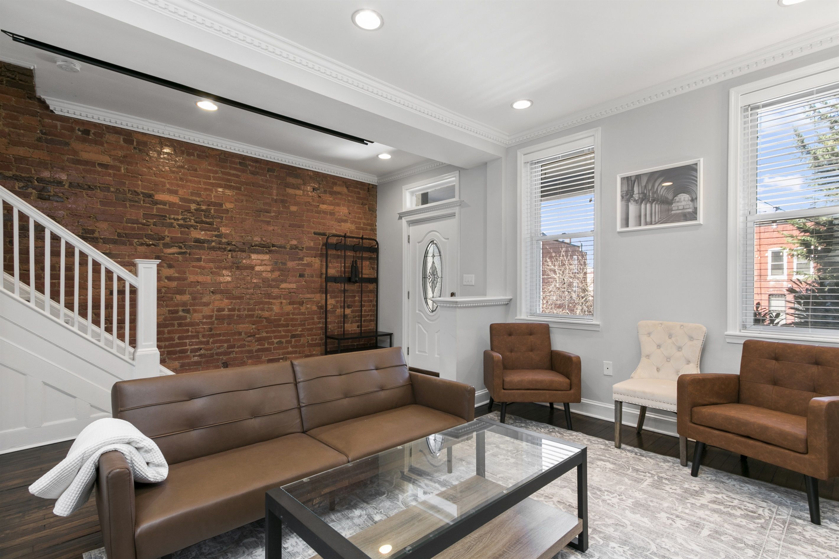 Un salon avec un canapé en cuir marron, deux fauteuils marron, une table basse en verre et un fauteuil blanc. On peut voir un mur en briques et une porte blanche.