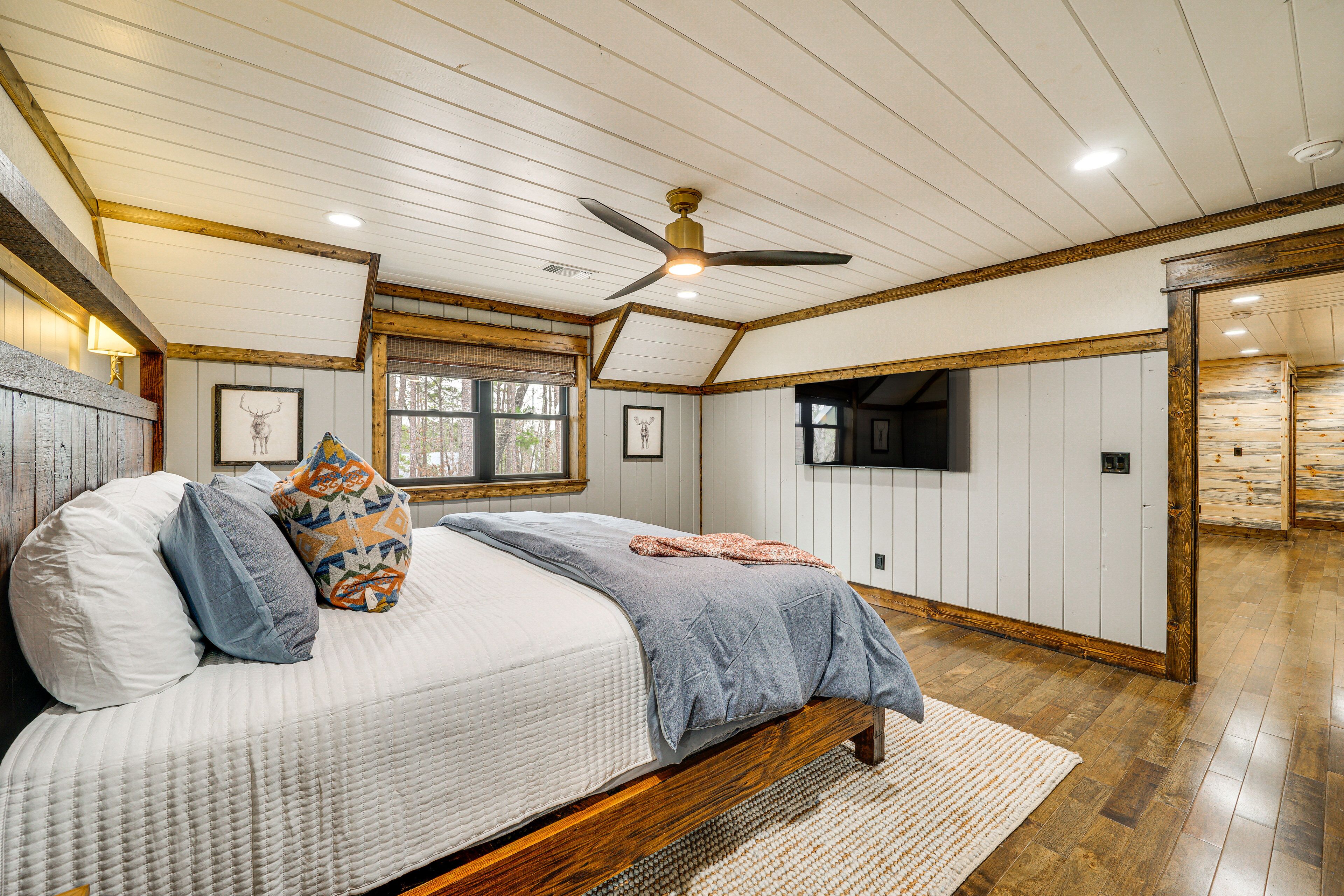 A bedroom with a bed, pillows, a ceiling fan, wooden beams, a window, and a TV.