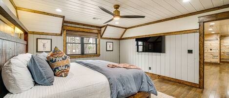 A bedroom with a bed, pillows, a ceiling fan, wooden beams, a window, and a TV.