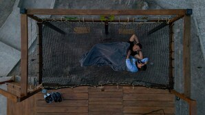 A hammock suspended between wooden beams, with a pair of shoes and a metal basket on the floor below.