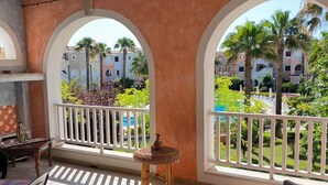 Un balcon donnant sur une piscine et des palmiers, une chaise en osier et une petite table sur laquelle est posé un vase.