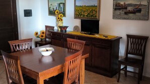 A dining area with a wooden table and chairs, a vase with sunflowers, and paintings on the wall. - BORGO SAN BASILIO APPARTAMENTO PENELOPE-CON PISCINA E ANIMAZIONE DIURNA E SERALE (Pisticci)
