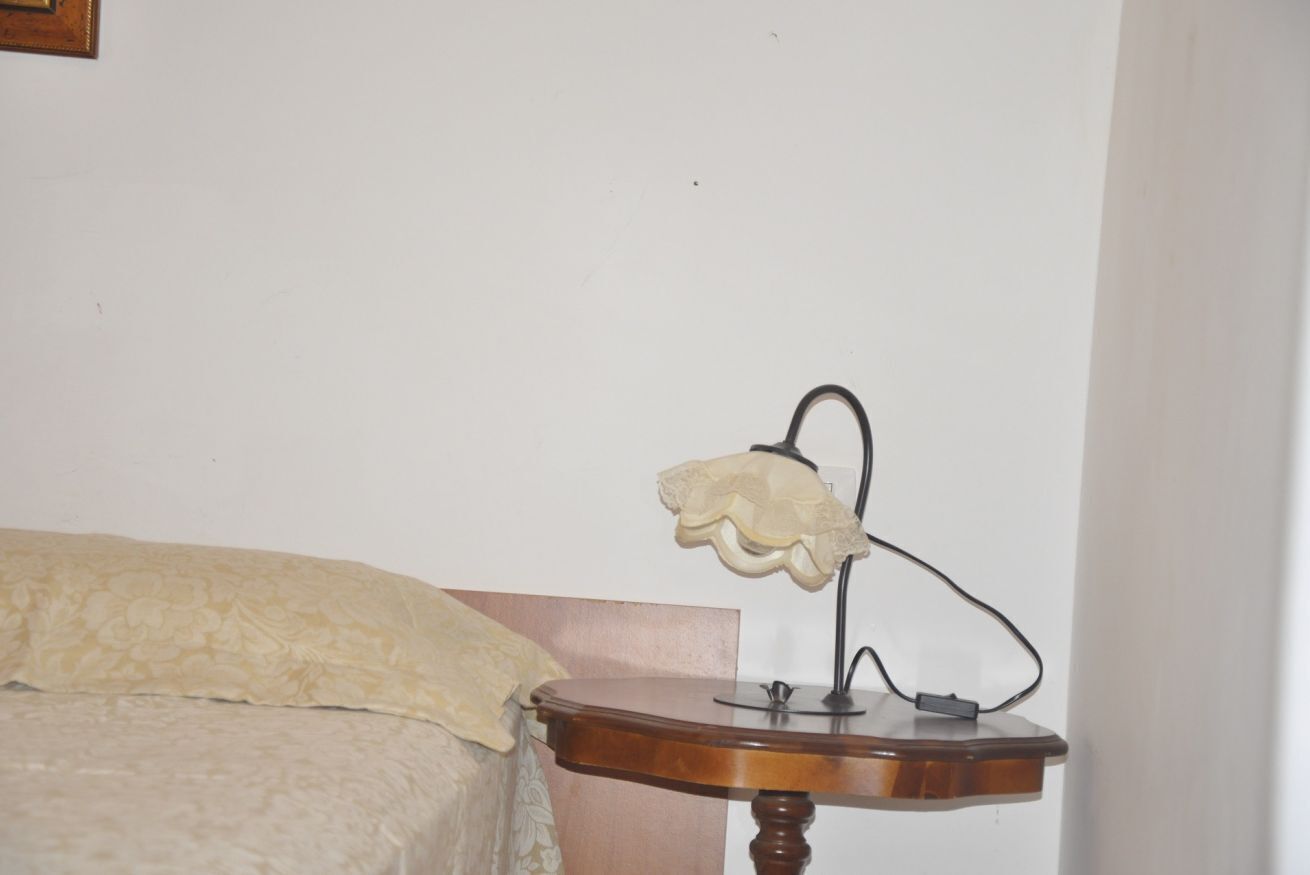 A bed with a beige patterned bedspread, a wooden bedside table with a lamp, and a wall with a framed picture.
