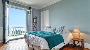 A bedroom with a large bed, a nightstand, a lamp, and a view of the sea through an open door.