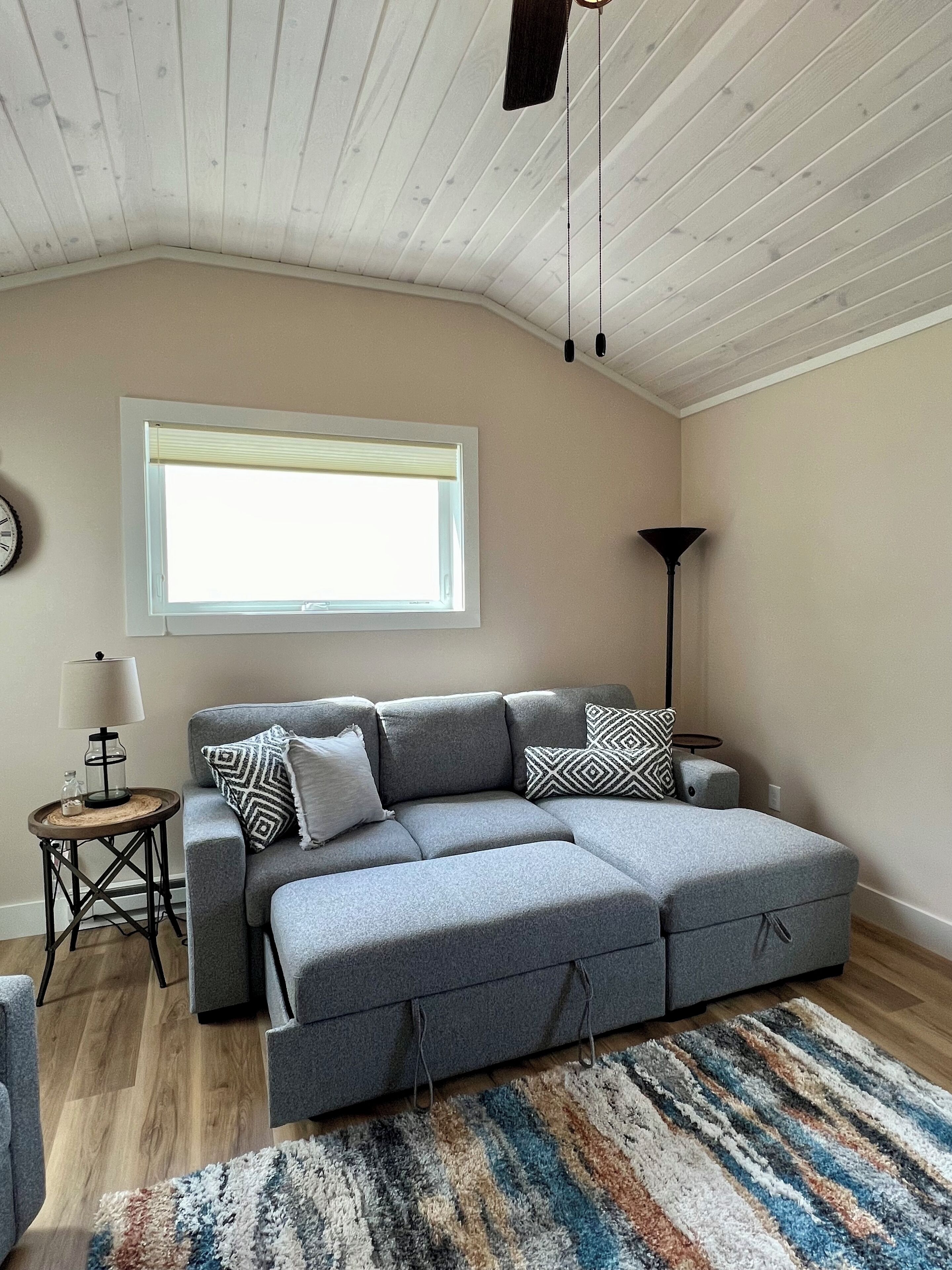 Un salon moderne avec un canapé d’angle gris, une petite table ronde avec une lampe, un lampadaire, une grande fenêtre et un tapis à motifs.
