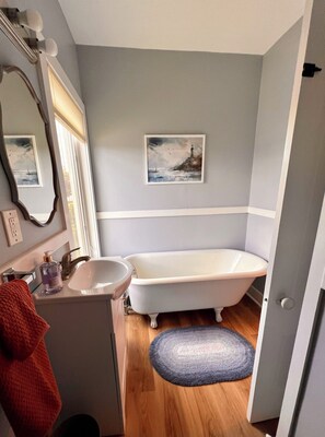 A bathroom with a white bathtub, a sink, and a mirror. There is a painting on the wall and a rug on the floor.