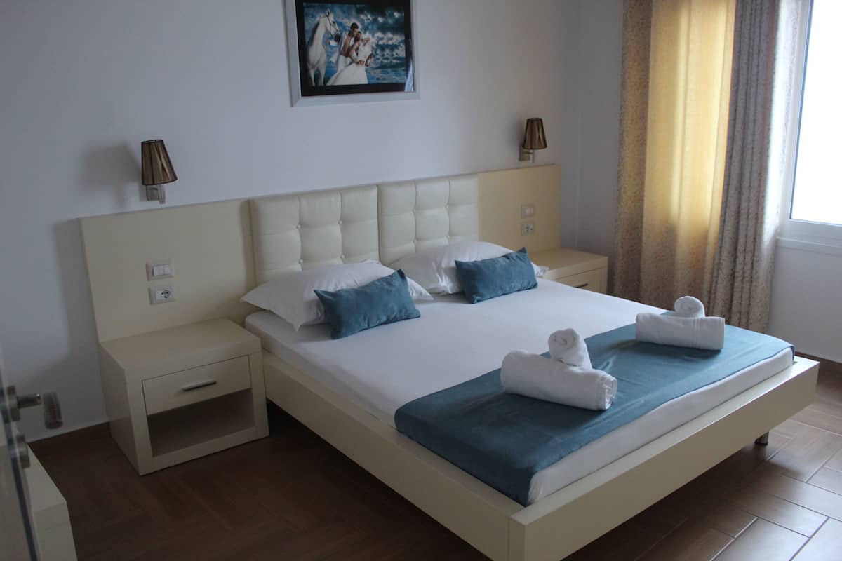 A large bed with a blue blanket and two pillows, a bedside table, a nightstand, and a wall-mounted lamp.
