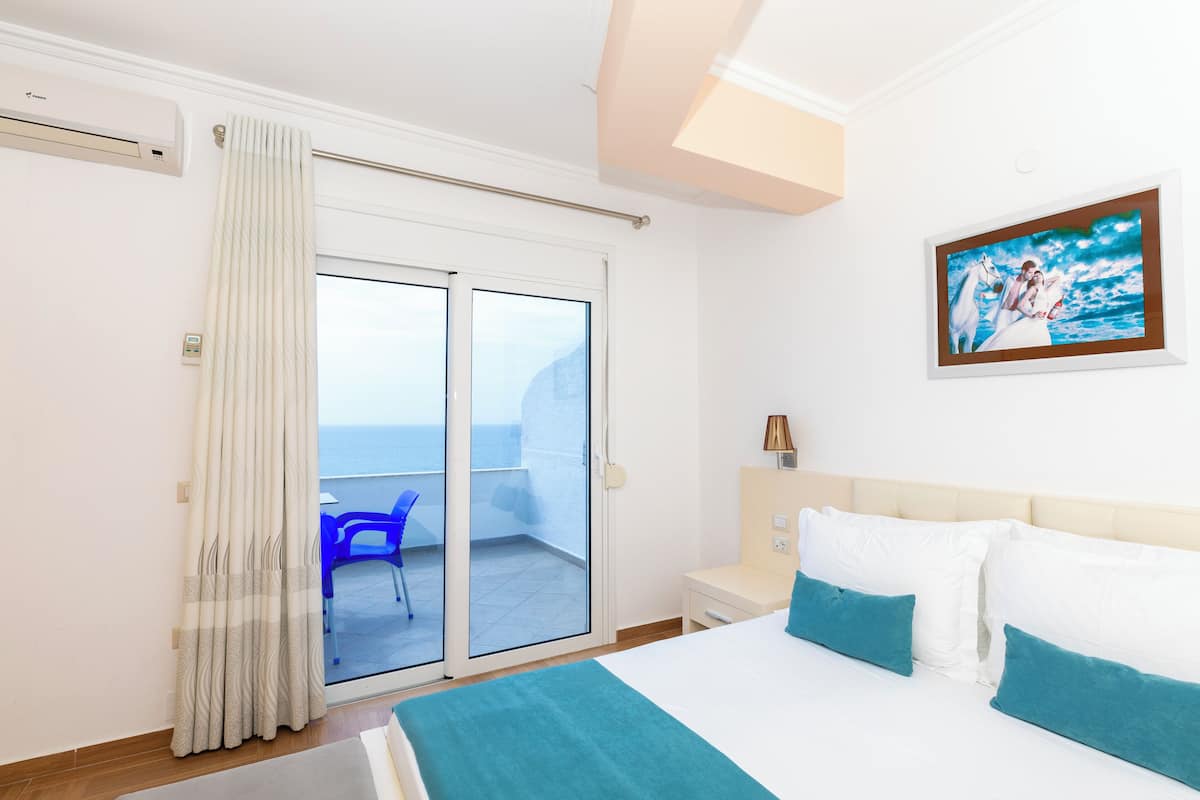 A hotel room with a bed, a nightstand, a painting, a view of the sea through a sliding door, and a blue chair outside.