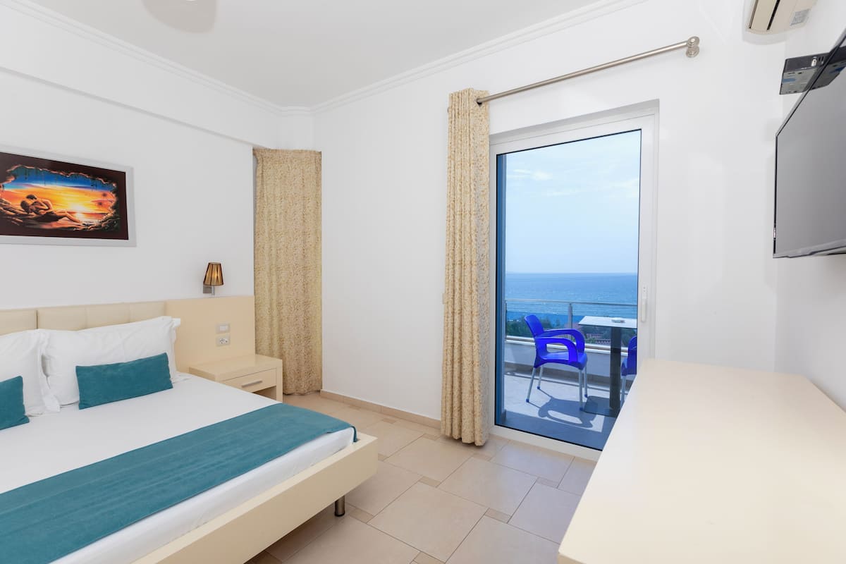 A hotel room with a bed, a desk, a chair, a painting, and a view of the sea through a large window.
