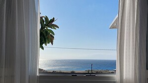 A window with sheer curtains, a view of the ocean, and a palm tree. - Venus - Venus Myconian Residences (Cyclades)