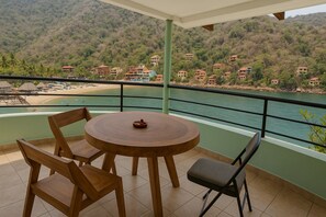 Un balcon doté d’une table ronde en bois et de chaises, offrant une vue sur une plage et une colline parsemée de bâtiments.