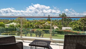 Ein Balkon mit Korbmöbeln, einer Glasreling und Blick auf Meer und Grünflächen.