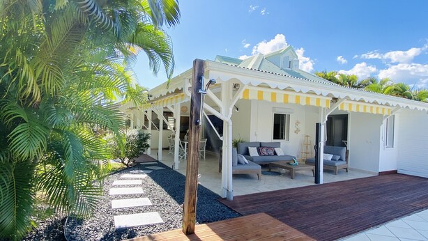 Ein überdachter Außenbereich mit Holzterrasse, ein weißes Gebäude mit gelber Streuung und eine Palme.