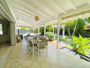 Un patio techado con una mesa y sillas, una lámpara colgante y vistas a una piscina y jardín.