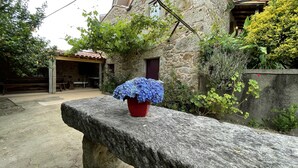 A stone bench with a potted blue hydrangea in front of a rustic stone building with a wooden door and a covered outdoor seating area.