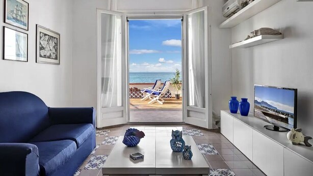 A living room with a view of the beach, a sofa, a coffee table with decorative items, and a TV on a white cabinet. - Raffaela's House (Massa Lubrense)