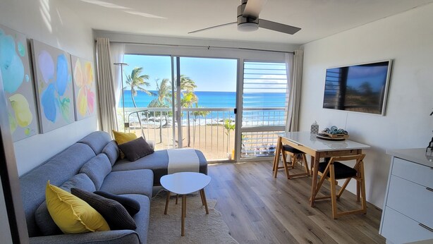 Un soggiorno moderno con un divano grigio, un piccolo tavolino bianco e pavimento in legno. È presente un balcone con vista sull'oceano e palme.