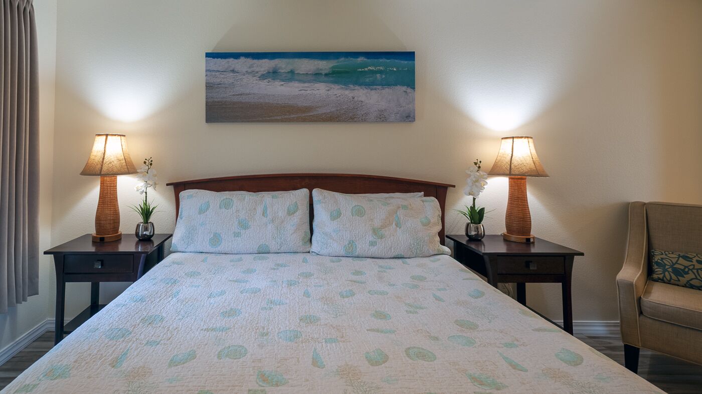 Une chambre d’hôtel avec un grand lit, deux tables de chevet avec des lampes, une chaise et un tableau représentant une scène de plage accroché au mur.