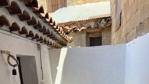Um pequeno pátio externo com duas cadeiras de madeira, uma mesa redonda, paredes brancas e chão em azulejo.