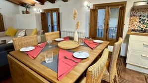 Uma sala de jantar com uma mesa de madeira posta para quatro pessoas, cadeiras de vime e vista para o exterior através de uma janela.