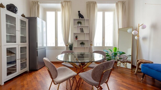 A dining area with a glass table, four chairs, a cabinet, a mirror, and a blue sofa.
