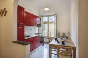 A compact kitchen with red cabinets, a dining table with chairs, and a window with a view of buildings. - Palazzo Borghese 2 Bedroom (Firenze)