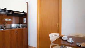 A compact kitchen with wooden cabinets, a stainless steel sink, and a small countertop. There is a wooden door with a brass handle and a white chair at a wooden table with a plate of croissants and cups. - Apartment by the sea with swimming pool (Porto Empedocle)