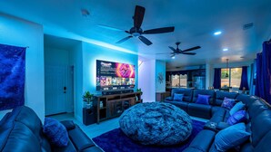 Ein modernes Wohnzimmer mit großem Flachbildfernseher, bequemen Sitzmöglichkeiten, Deckenventilatoren und Blick in die Küche.