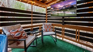 Een overdekt terras met een televisiescherm dat een berglandschap toont, houten balken en een vloer van groen kunstgras.