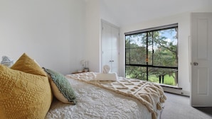 Un dormitorio con un ventanal grande, una cama con edredón blanco y vistas al verdor exterior.