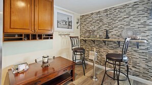 A kitchen with wooden cabinets, a dining table, two bar stools, a decorative wall with a cityscape picture, and a shelf with kitchenware. - C & N Lodging, Bluffton, SC (Bluffton)